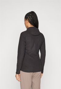 Woman with braided hair wearing a black textured hooded jacket and beige pants, facing away against a plain white background.