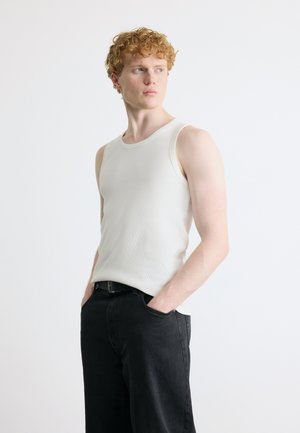 Junger Mann mit lockigem rotem Haar, der ein weißes ärmelloses Hemd und schwarze Hose trägt, mit den Händen in den Taschen stehend und zur Seite schauend.