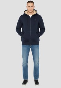 Marineblauwe hoodie met rits en een capuchon met bontvoering, kangeroezakken en geribbelde boorden. Combinatie met lichtblauwe spijkerbroek en zwarte sneakers.