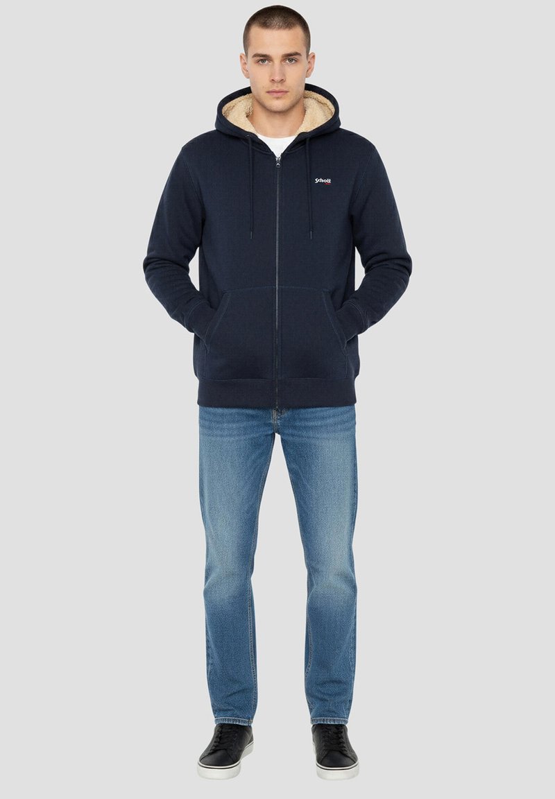 Marineblauwe hoodie met rits en een capuchon met bontvoering, kangeroezakken en geribbelde boorden. Combinatie met lichtblauwe spijkerbroek en zwarte sneakers.