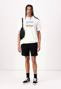 Hvid t-shirt med et farverigt grafisk print, sorte shorts og sorte sneakers. Sort crossbody taske med justerbar rem.