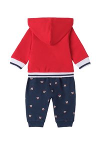 Czerwona bluza z zamkiem błyskawicznym i niebieskimi spodniami w wzór misia. Bluza ma białe i granatowe paski na mankietach i dolnym obszyciu oraz kaptur.