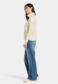 Krämfärgad fleecepullover med hög krage, med en texturerad yta och enfärgade ärmar, matchad med löst skurna blå jeans.