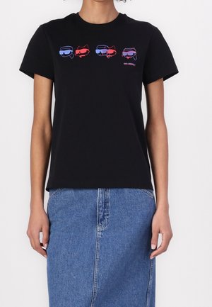Person trägt ein schwarzes T-Shirt mit blauen und roten Comic-Gesichtern mit Sonnenbrille und einem blauen Jeansrock.