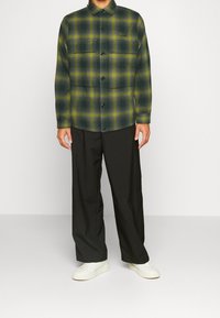 Grön och gul rutigt skjorta med knappar, långärmad och två framfickor, kombinerad med svarta byxor och vita sneakers.