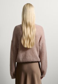 Maglione lavorato a maglia rosa chiaro con vestibilità ampia, tessuto testurizzato e maniche lunghe, abbinato a una gonna marrone liscia. Capelli biondi lunghi e lisci visibili.