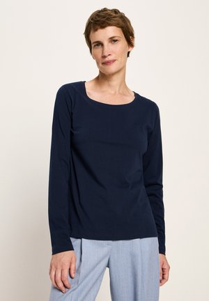 Frau mit kurzen braunen Haaren, die ein marineblaues Langarmoberteil und hellgraue Hose trägt und vor einem schlichten hellen Hintergrund steht.