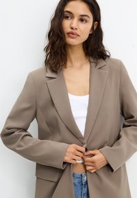 Blazer beige avec un design structuré, présentant des revers larges, une fermeture à un bouton et des manches longues. Porté par-dessus un top blanc.