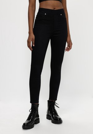 Frau trägt schwarze Skinny-Jeans mit hoher Taille und glänzende schwarze Schnür-Combat-Stiefel, steht vor einem weißen Hintergrund.