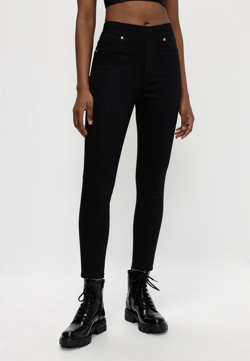 Femme portant un jean skinny noir taille haute et des bottes de combat noires brillantes à lacets, debout sur un fond blanc uni.