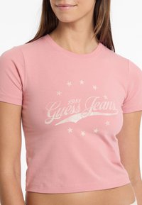 Rožinė trumpa rankovė T-marškiniai su balta "Guess Jeans" grafika, žvaigždėmis ir metų "1981" užrašu, pagaminti iš švelnios medvilnės.
