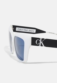 Lunettes de soleil blanches surdimensionnées avec des verres bleus. Présentent une finition mate et une branche noire avec le logo "Calvin Klein Jeans" et des détails d'accent.