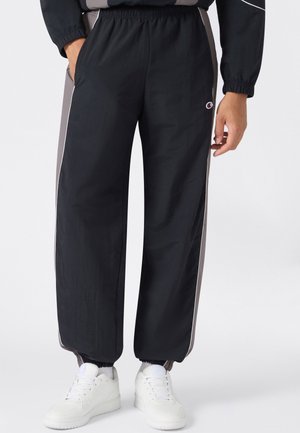 Person trägt schwarze und graue Jogginghosen mit elastischen Bündchen und weiße Sneakers, steht mit einer Hand in der Tasche.