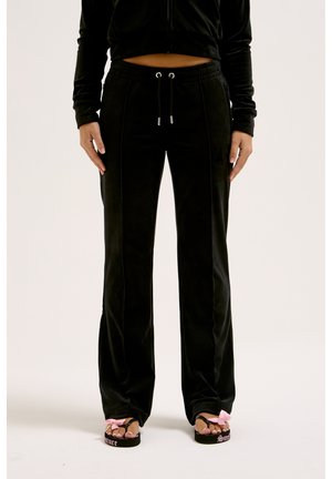 Person iført sorte velourbukser med snøre i taljen og sorte flip-flops med lyserøde sløjfer på tæerne står mod en lys baggrund.