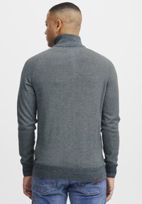 Fitted dark grey sweater with a high collar, ribbed cuffs, and hem. Knit texture with subtle patterning, paired with blue jeans.