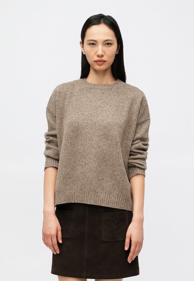Jeune femme aux longs cheveux noirs portant un pull en laine marron et une jupe marron foncé, debout devant un fond blanc uni.