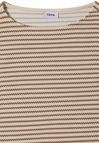 Gros plan d'un tissu tricoté beige et marron à rayures horizontales avec de petits carrés texturés et une étiquette blanche portant la mention "CECIL" cousue à l'encolure.