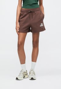 Καφέ βαμβακερά σορτς με ελαστική μέση και κορδόνι, με εκτύπωση λευκού λογότυπου. Συνδυάζονται με μπεζ αθλητικά παπούτσια και λευκές κάλτσες μέσης.