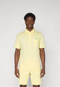 Cămașă polo galben deschis cu guler, având un buzunar frontal și un logo verde. Asortată cu pantaloni scurți în aceeași nuanță, ambele dintr-un material neted.