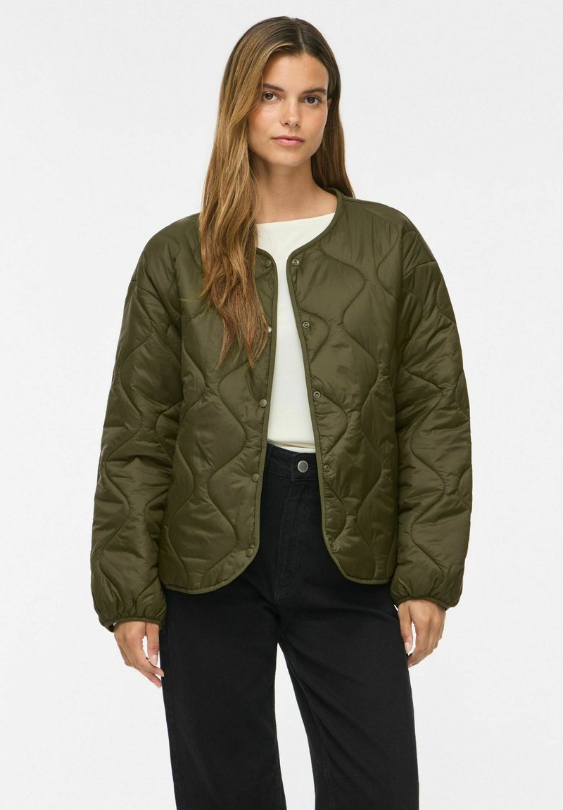 Femme aux longs cheveux bruns portant une veste matelassée vert olive sur un haut blanc et un pantalon noir, debout devant un fond uni.