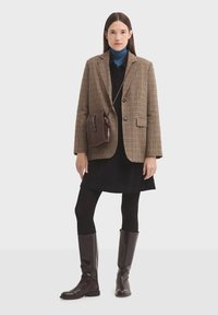 Hnedý kockovaný blejzer cez čierne šaty, spárovaný s čiernymi pančuchami a hnedými vysokými čižmami. Malá hnedá kabelka cez plece dotvára celý outfit.