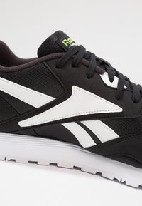 Chaussure de sport noire avec des accents blancs. Elle présente une partie supérieure en tissu texturé, un col rembourré et une semelle blanche contrastante avec un motif antidérapant.