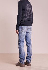 Černé tričko s dlouhým rukávem s motivem lebky, modré oprané džíny s detaily šití a hnědá obuv.