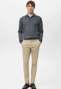 Młody mężczyzna stojący z rękami w kieszeniach, ubrany w ciemnoszarą koszulkę polo z długim rękawem, beżowe spodnie i czarne buty na jednolitym białym tle.