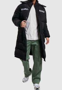 Svart pufferjacka med "KARL" och "KANI" logotyper, dragkedja fram, huva och elastiska ärmslut; matchad med gröna cargobyxor och silversneakers.