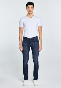 Młody mężczyzna stojący, ubrany w białą koszulkę z dekoltem w serek, ciemnoniebieskie dżinsy i białe trampki, na tle jasnego, jednolitego tła.