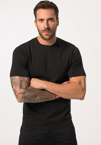 Černé tričko s krátkým rukávem vyrobené z hladké tkaniny s kulatým výstřihem. Obsahuje složité tetování na pažích, žádné viditelné logo nebo vzory.