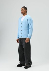 Cardigan bleu clair en maille avec col en V, cinq boutons et deux poches avant ; associé à un jean noir ample et des chaussures noires.