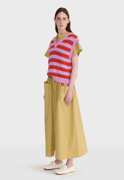 Jeune femme aux longs cheveux bruns portant un pull sans manches à rayures roses et rouges par-dessus une robe jaune moutarde avec de grandes poches et des chaussures blanches.