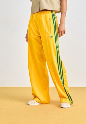 Personne portant un pantalon de survêtement Adidas jaune avec des bandes vertes et des baskets beiges, debout sur un sol jaune devant un fond uni.