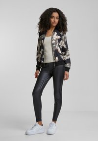Veste bomber noire avec des motifs de feuilles blancs et beiges, associée à des leggings noirs brillants et des baskets blanches. Texture lisse et design ajusté.