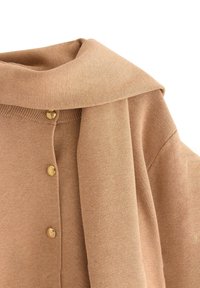 Beige neulepusero, jossa on leveä kaulus ja kolme kultanappia edessä, pehmeä tekstuuri ja laskeutuva muotoilu.