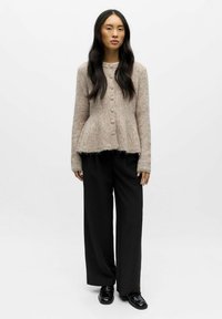 Lysebrun, tekstureret cardigan med knapper foran, kombineret med brede sorte bukser. Blødt stof og detaljer med frynset kant synlige.