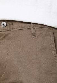 Pantalon en coton marron avec une texture lisse, doté d'un bouton métallique et d'une passants de ceinture à la taille, partiellement rentré sous un haut blanc.