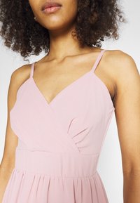 Světle růžové, bezramenné šaty s překládaným designem, s tenkými ramínky a nařaseným pasem, vyrobené z hladkého materiálu s jemnou texturou.