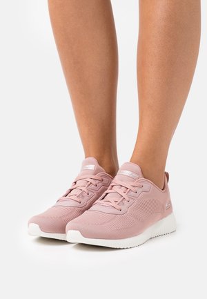 Jambes portant des baskets Skechers rose clair avec des semelles blanches, debout sur une surface blanche unie.