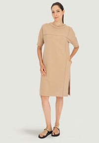 Beżowa sukienka do kolan z krótkimi rękawami, okrągłym dekoltem, bocznymi rozporkami i subtelnymi detalami zszycia. Noszona z czarnymi sandałami.
