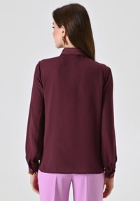 Blusa bordeaux a maniche lunghe con colletto, texture liscia e dettagli con polsino elasticizzato, abbinata a pantaloni rosa chiaro.