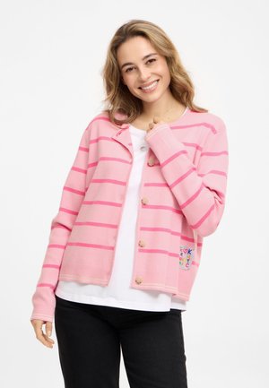 Lächelnde Frau mit blondem Haar trägt eine rosa gestreifte, geknöpfte Strickjacke über einem weißen Hemd und schwarzen Hosen vor weißem Hintergrund.