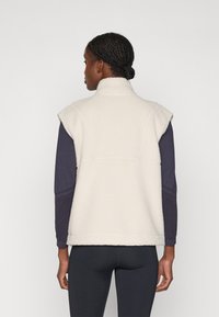 Fleece vest in crème met een hoge kraag en een mouwloos ontwerp. Heeft een gestructureerd, zacht oppervlak en een contrasterend donker langärmige top eronder.