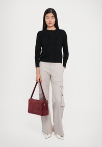 Nainen seisoo päällä musta napillinen neuletakki, beige cargo-housut, valkoiset kärkipohjat ja pidä punainen käsilaukku sivullaan.