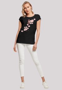 Black t-shirt with short sleeves, featuring pink cherry blossom graphics and Japanese text on the left side; smooth cotton fabric.