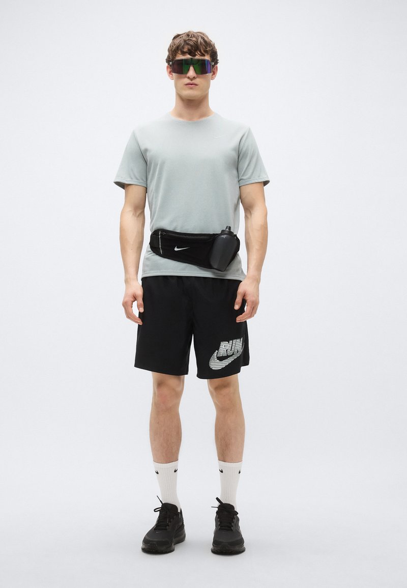Graues T-Shirt mit kurzen Ärmeln, schwarze Shorts mit "RUN"-Grafik, schwarze Bauchtasche, weiße Socken mit schwarzen Akzenten, dunkle Turnschuhe und reflektierende Sonnenbrille.