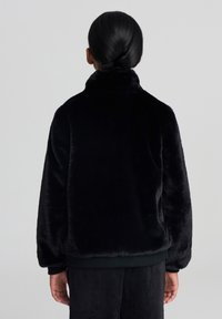 Giacca nera in pelliccia sintetica con collare alto, caratterizzata da una texture morbida e polsini elasticizzati, vista dal retro.