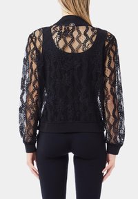 Giacca bomber nera in pizzo con motivi floreali, maniche lunghe e trasparenti, e una vita nera solida. Presenta un colletto rotondo sul retro e un dettaglio con logo.