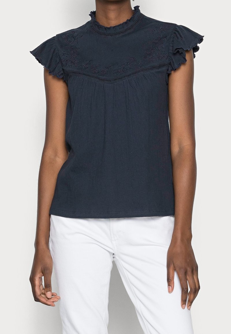 Femme portant un chemisier sans manches bleu marine foncé avec un bord volanté et des détails floraux brodés, associé à un pantalon blanc.